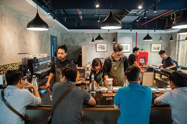 Baristas and customers interact at a modern coffee shop counter with hanging lights.