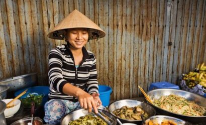 Woman in conical hat serving food from bowls at a market stall.
