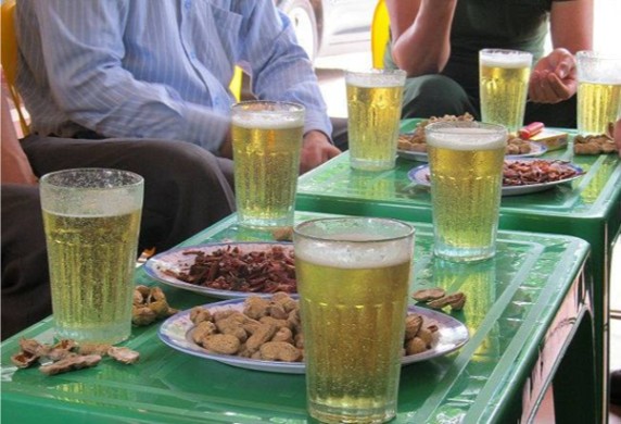 Glasses of beer and plates of snacks on green tables with people sitting nearby.