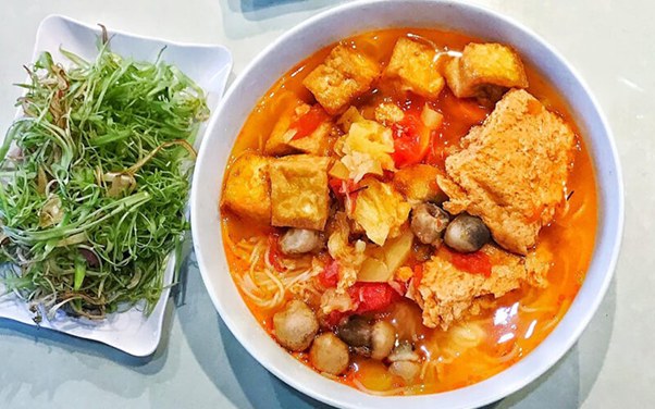 Bowl of noodle soup with vegetables, tofu, and herbs beside a plate of greens.