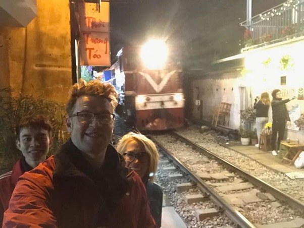 Group takes selfie near train on narrow street at night.