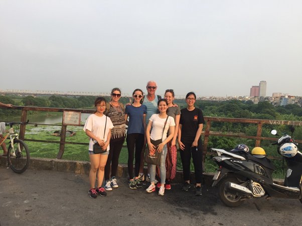 Group of seven people posing on a bridge with greenery and city skyline in the background.