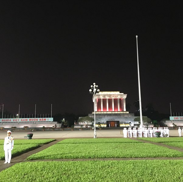 Lit building with columns at night, surrounded by soldiers in white uniforms and green grass in the foreground.