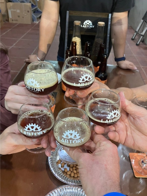 Five people clinking Labtory beer glasses over a wooden table with snacks.
