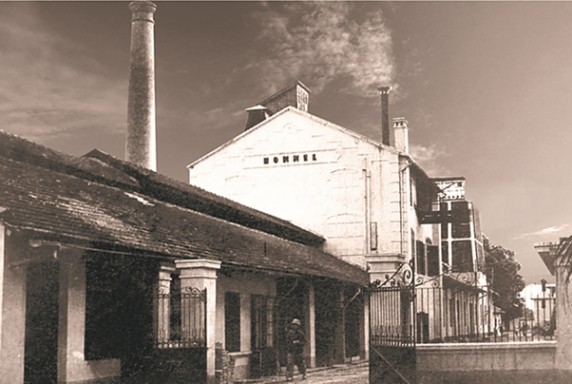 Sepia-toned image of an old factory building with smokestack and people.